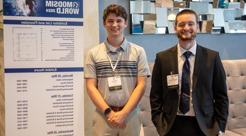 Two young men stand in front of a conference sign.